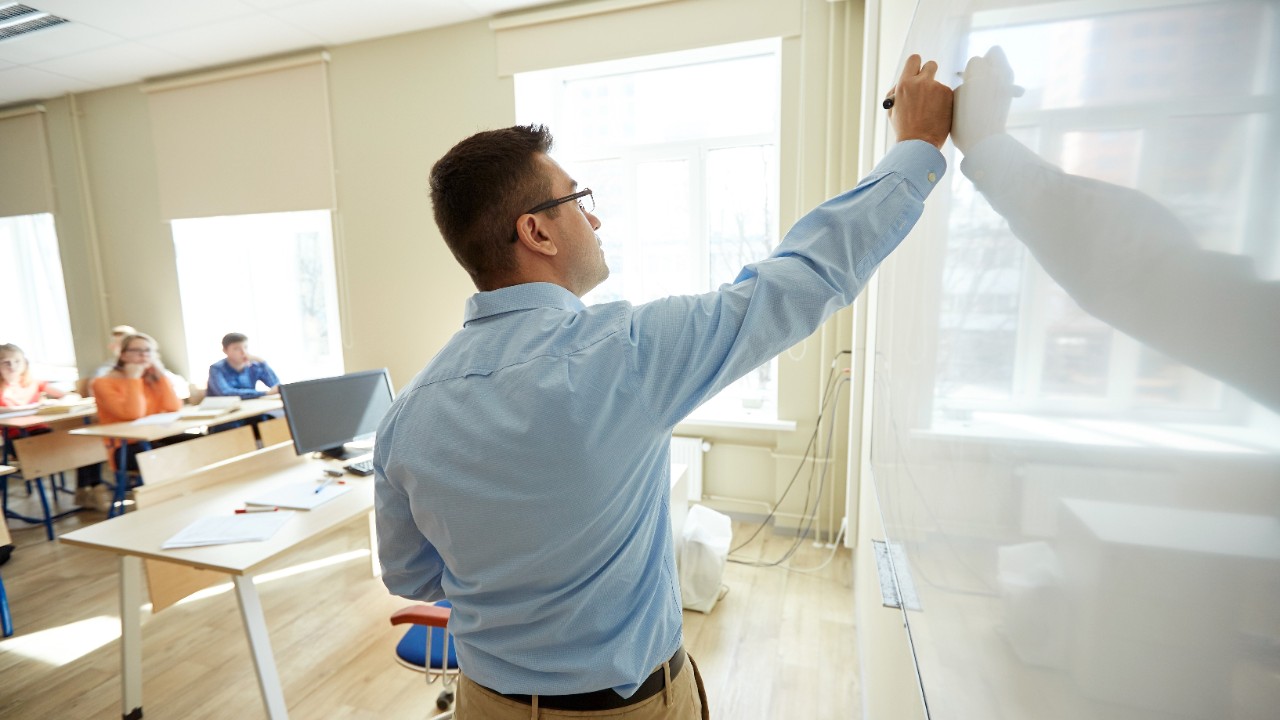 education, school, learning, teaching and people concept - teacher standing in front of students and writing something on white board in classroom