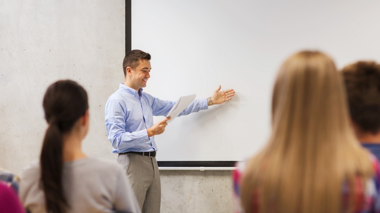 education, high school, technology and people concept - smiling teacher with notepad, laptop computer standing in front of students and showing something on white board in classroom