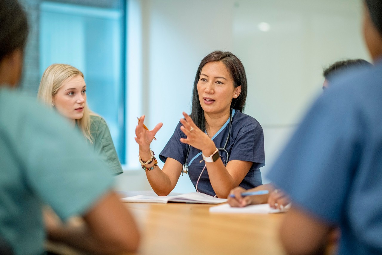 A small group of Nurse sit at a boardroom table as they meet to discuss patient cases.  They are each dressed professionally in scrubs and  have stethoscopes around their necks as they discuss plans of care for each case.